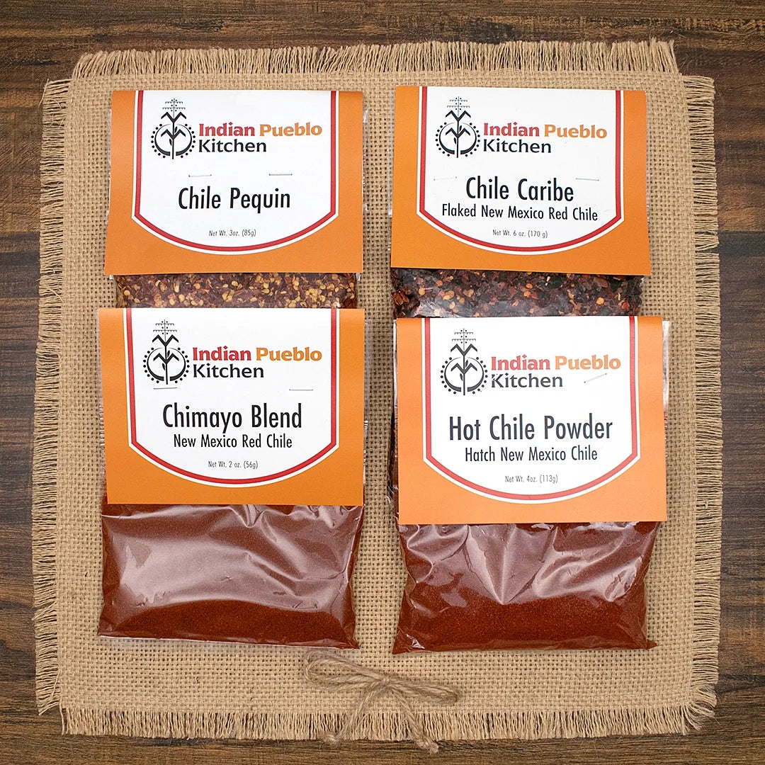 Four bags of Indian Pueblo Kitchen chile powder arranged on a burlap mat over a dark wood surface. The packages include Chile Pequin, Chile Caribe, Chimayo Blend, and Hatch New Mexico Hot Chile Powder.